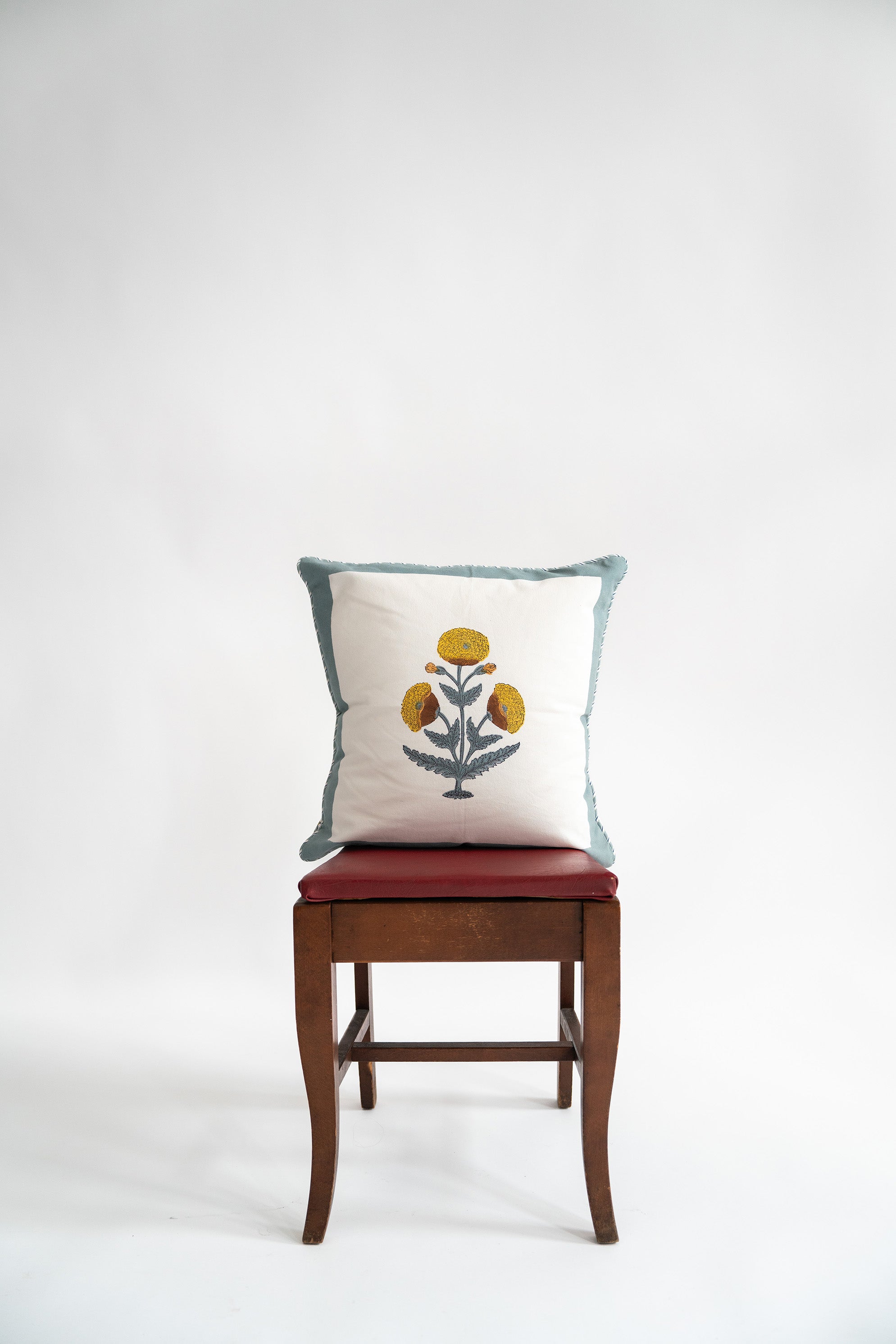 Blue poppy blockprint decorative pillow with yellow floral design on white fabric and blue trim, displayed on wooden chair
