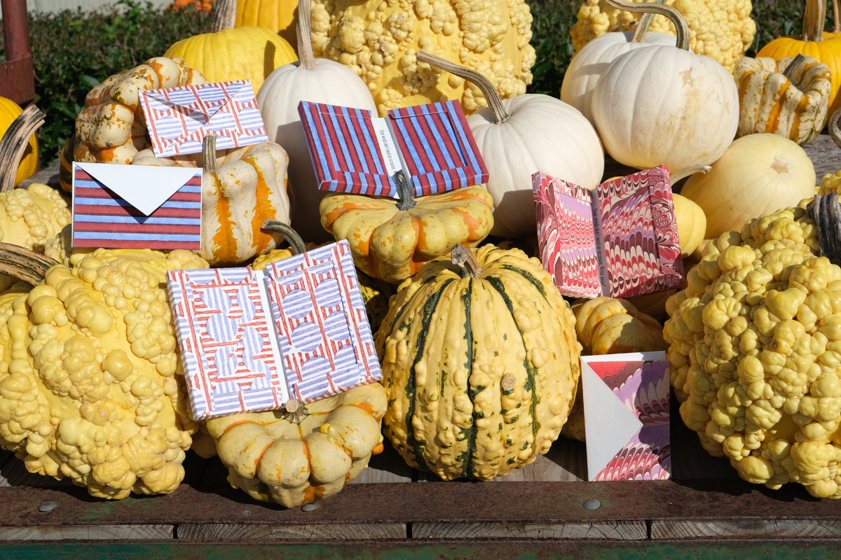 A collection of decorative gift boxes wrapped in colorful patterned paper and adorned with white envelopes, arranged among various yellow, orange, and white pumpkins on a wooden surface.